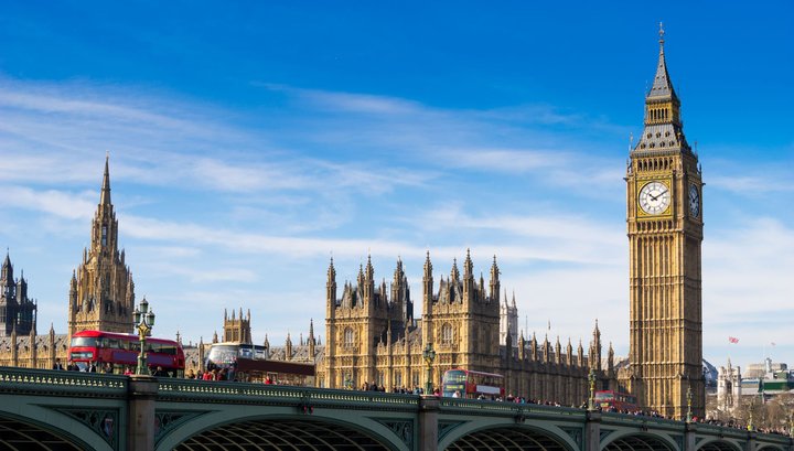 View of the Palace of Westminster and Big Ben in London, with red double-decker buses crossing Westminster Bridge under a bright blue sky.