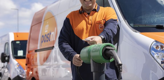 Smiling delivery driver in a uniform standing in front of an electric van, holding a charging cable and preparing to charge the vehicle.