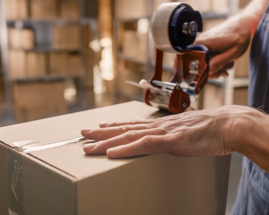 Close-up of a worker sealing a cardboard box with packing tape in a warehouse filled with shelves and packages.