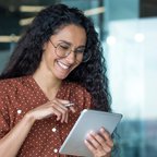 Smiling woman with curly hair and glasses working in a modern office, using a tablet and stylus while seated at a desk with a laptop and papers.