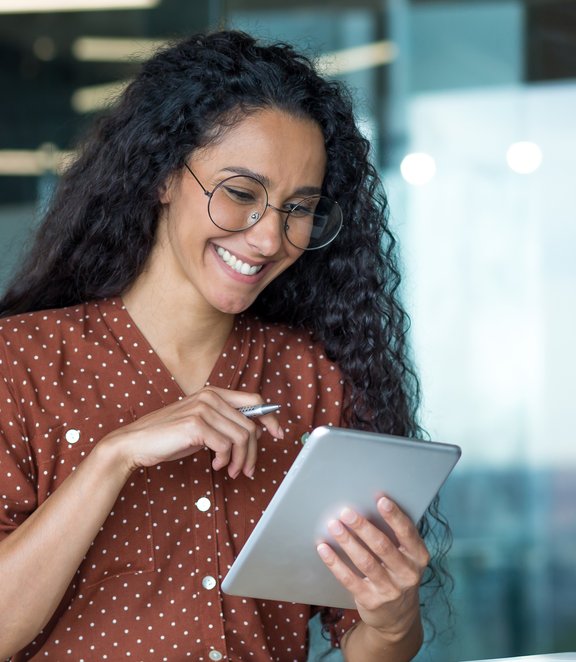Smiling woman with curly hair and glasses working in a modern office, using a tablet and stylus while seated at a desk with a laptop and papers.